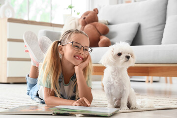 Fototapeta premium Cute little happy girl with Maltese dog reading book on floor at home