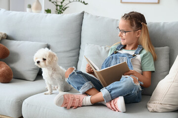 Fototapeta premium Cute little happy girl with Maltese dog reading book on sofa at home