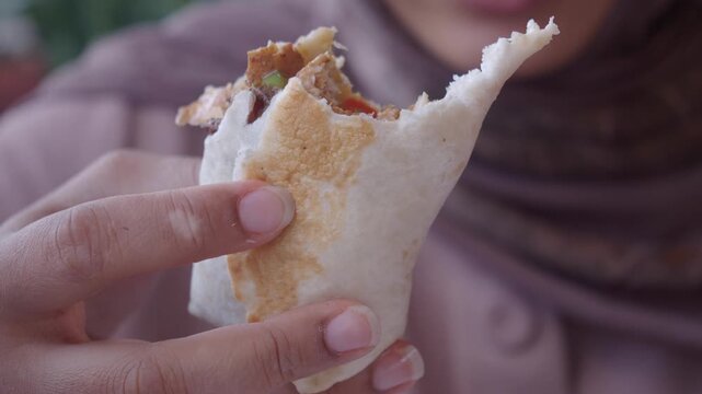 Close up of person in hijab eating shawarma wrap with chicken and vegetables