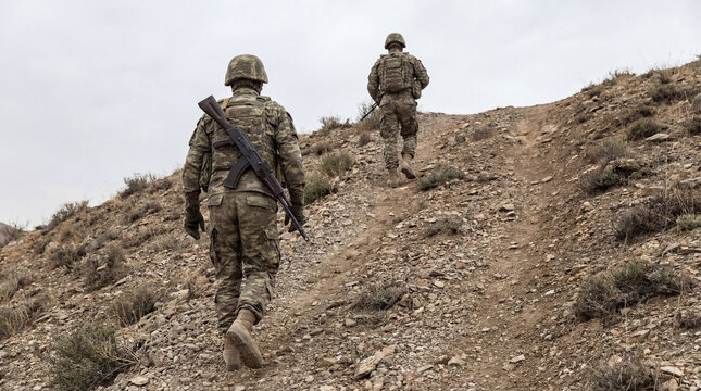 Two armed soldiers in military camouflage climb a barren, rocky desert landscape.