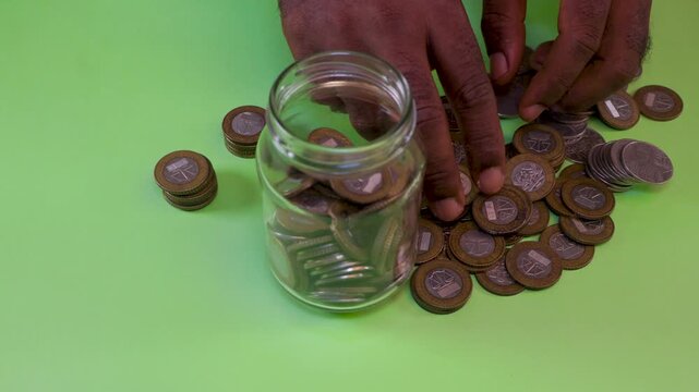 Hands sorting and placing two-toned Ghanaian Cedi coins into a glass jar over a green surface. A visual metaphor for savings, personal finance, and accumulating wealth through small contributions.