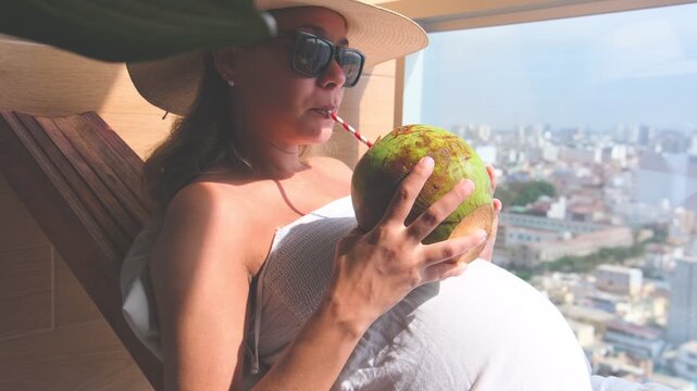 Beautiful young woman relaxing in a sun lounger drinking from a fresh green coconut on a summer day