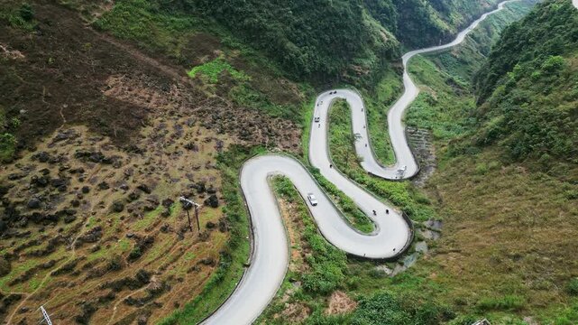 Aerial view of a serpentine mountain road with tight curves and sweeping bends crossing steep green hills as vehicles move through a dramatic landscape at Ma Pi Leng Pass in Ha Giang.