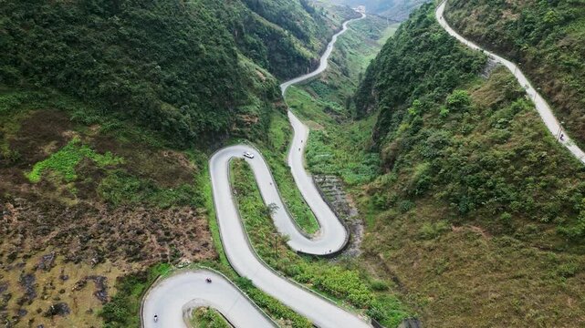Aerial shot captures a high mountain road with flowing curves and sharp bends crossing steep hills with vehicles traveling through a vast and rugged landscape in Ha Giang northern Vietnam