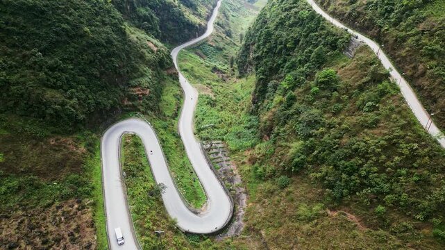 Aerial drone footage captures a winding mountain pass with sharp hairpin turns cutting through steep green hills as motorbikes and cars travel along the iconic Ma Pi Leng Pass in northern Vietnam