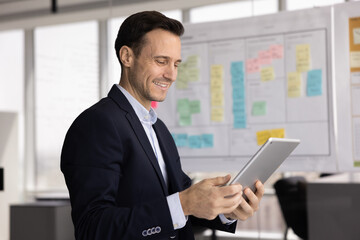 Fototapeta premium Digital planning and analysis. Smiling modern mid aged businessman wearing formal suit use electronic pad device app in work office near agile task board review analytics online optimize project tasks