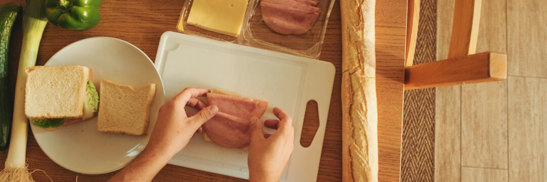 Close-up of the hands of guy dressed in denim shirt cooking sandwich while standing in the kitchen, View from above. Panoramic