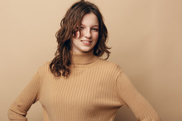 Smiling young woman with curly brown hair wearing a beige knitted sweater stands confidently against a plain beige background. Concept of casual style and natural beauty. © SHOTPRIME STUDIO