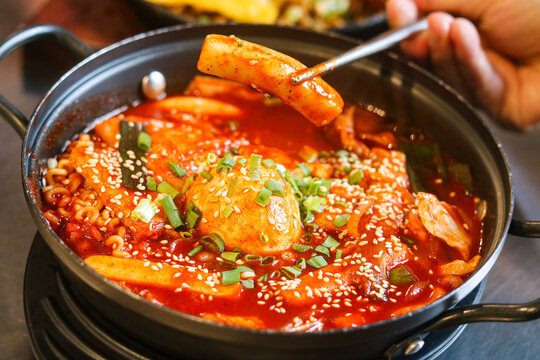 Eating a serving of Tteokbokki with chopsticks. This korean dish features rice cakes (tteok), fish cakes (eomuk), fresh green onions, and an egg, all coated in a red gochujang sauce.