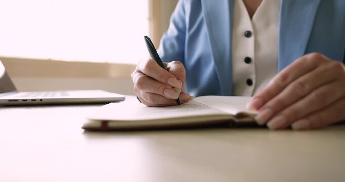 Hands of business professional woman writing notes in notebook, planning tasks for day, making schedule, working at laptop computer, making summary of online lecture, webinar. Cropped shot close up