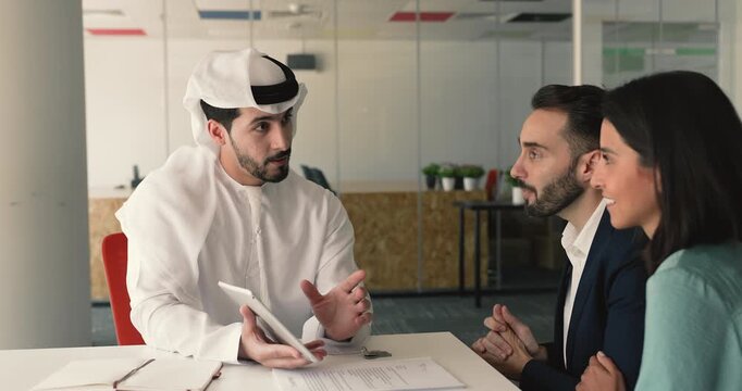 Happy young Caucasian couple consulting male Arabic real estate seller, watching virtual tour, property offer on tablet, discussing buying or rent legal terms, mortgage, investment