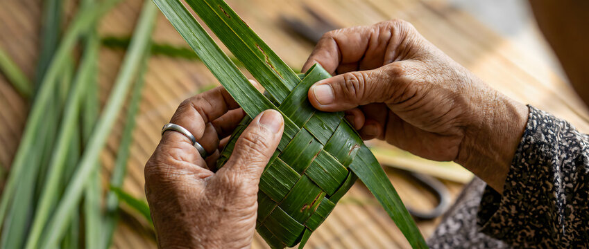 Close-up of an unrecognizable older person's hands skillfully weaving green palm leaves to make traditional ketupat