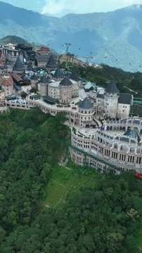 Vertical video, Aerial view of French Village in Da Nang, Vietnam, with castle architecture on mountain peak, surrounded by lush forest and cable car system, popular tourist destination.