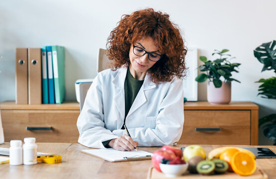 Beautiful nutritionist writing some notes while working with computer in the consultation