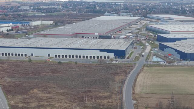 Wide aerial drone pan shot of a large modern logistics park with multiple warehouse buildings blue facades loading docks access roads and trucks visible across the complex in Poland