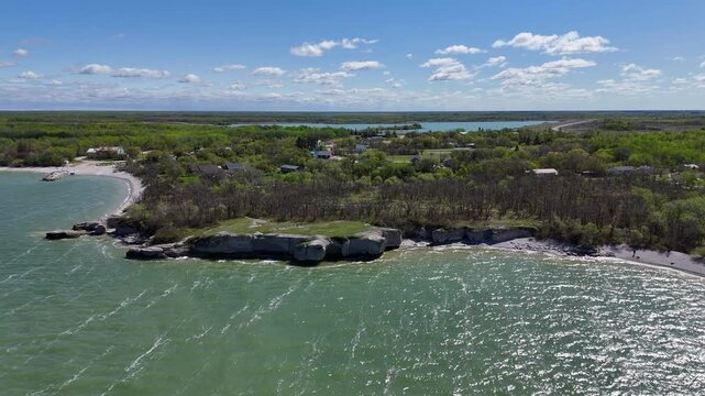 Drone shot farther out from limestone cliffs at Steep Rock Manitoba orbiting left, showing green water of Lake Manitoba, shoreline, and trees on a sunny summer day.