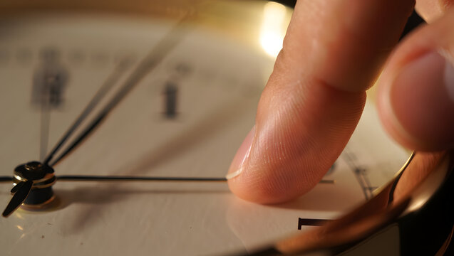 High-angle shot of a person's hand with the index finger touching the center of a metal-framed analog clock displaying a specific time on a light-colored surface