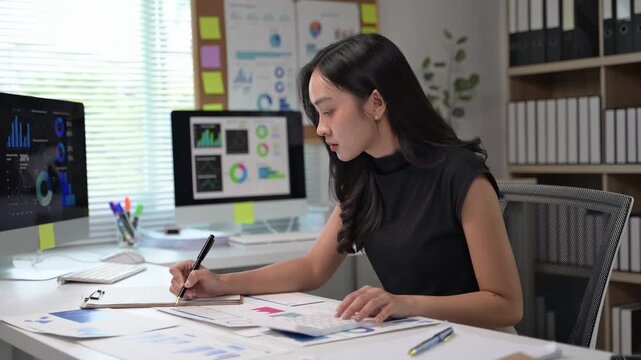 A woman is sitting at a desk with a computer monitor and a stack of papers. She is writing with a pen and she is focused on her work