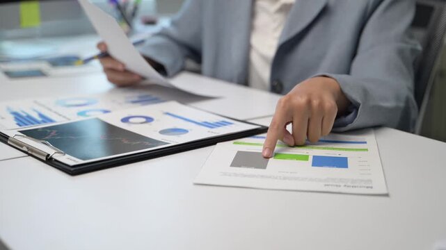 A woman is sitting at a desk with a stack of papers in front of her. She is pointing at a piece of paper with a finger. The papers are spread out on the desk
