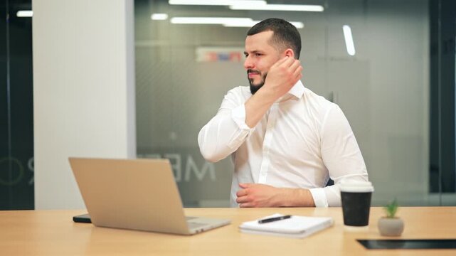 Tired man in white shirt itching hand due to allergy at office desk with laptop. Male executive suffering from skin irritation, dermatitis or itching during working day.