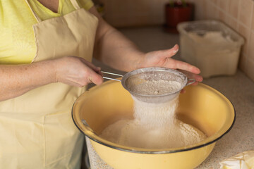 Fototapeta premium Adult woman hands sifting flour over bowl with sieve in kitchen