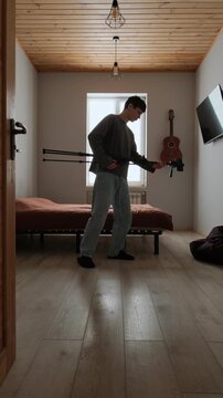Young man tired of filming in ordinary bedroom turns frustration into joke and plays air guitar with tripod. Content creator burnout with humor. Real life lifestyle behind the scenes. Vertical video