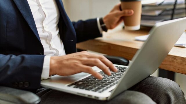 A professional man in a wheelchair works on his laptop while taking a sip of coffee in the office.