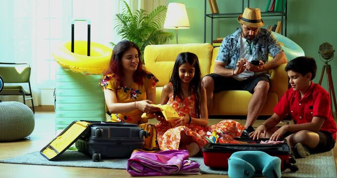 Happy Indian mother and kids playfully packing suitcases for a summer trip while the father sits on the sofa. Cheerful young family in tourist attire preparing for a holiday at home
