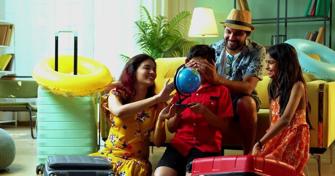 Cheerful Indian father closing his son's eyes as the boy puts a finger on a revolving globe to choose a travel destination. Happy Asian family planning a summer vacation trip at home