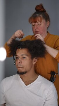Caucasian female stylist braiding the curly hair of a young african american man. Professional hairdresser creating a fashionable hairstyle