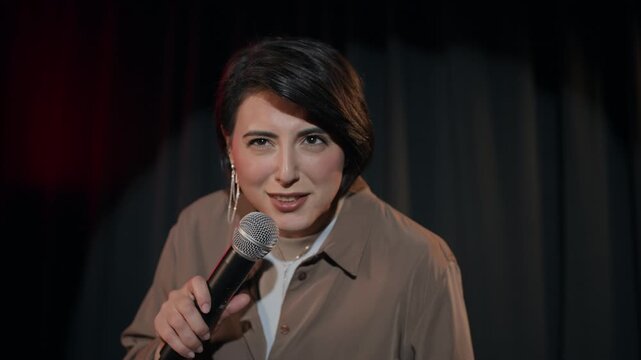 Medium shot of multiethnic young adult female comedian interacting with audience by asking questions during live stand up show on stage of comedy club, slow motion