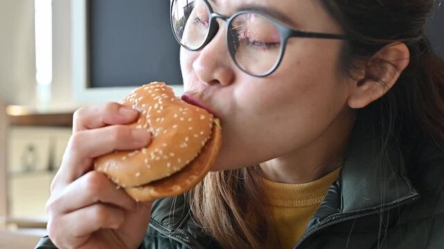 Footage of young Asian woman while eating a hamburger in cafe. A hamburger is a sandwich consisting of a cooked meat patty on a bun or roll.