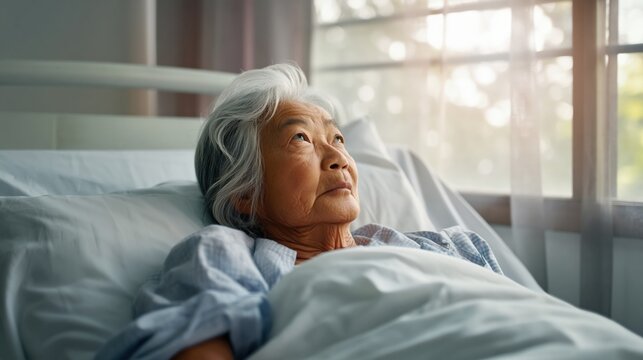 Elderly asian female patient lying in hospital bed gazing toward window light with peaceful contemplative expression showing resilience and calm in a quiet medical room at sunrise