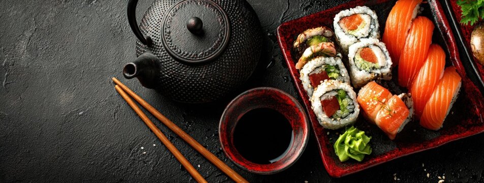Sushi platter featuring assorted rolls and sashimi with a black teapot and chopsticks on a dark textured surface, garnished with wasabi and soy sauce