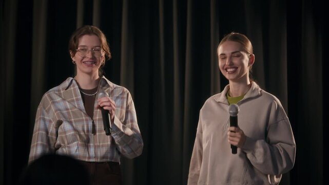 Medium shot of two cheerful female comedians giving high five while crowd working with audience during stand up show at comedy venue against black curtains