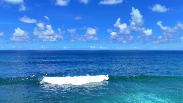 Aerial drone shot flying parallel to surfers riding waves near dramatic rocky cliffs in Bali, Indonesia. Dynamic action scene capturing the thrill of surfing in crystal clear ocean water