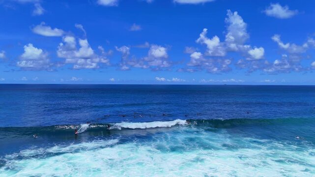 Aerial drone shot flying parallel to surfers riding waves near dramatic rocky cliffs in Bali, Indonesia. Dynamic action scene capturing the thrill of surfing in crystal clear ocean water