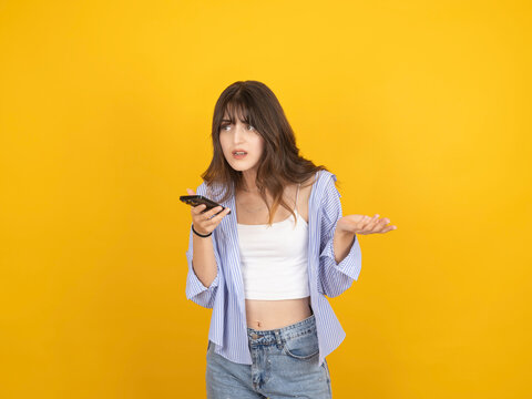 Confused young woman holding smartphone with questioning gesture, wearing striped shirt and denim, standing against bright yellow studio background with copy space. Problem and uncertainty concept.