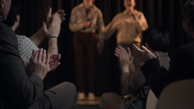 Shot from audience seats of crowd applauding to female stand up comedians bowing on stage at end of live show at comedy venue, copy space