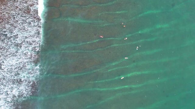 Aerial drone shot flying parallel to surfers riding waves near dramatic rocky cliffs in Bali, Indonesia. Dynamic action scene capturing the thrill of surfing in crystal clear ocean water