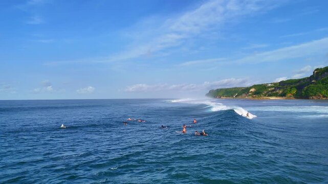 Aerial drone shot flying parallel to surfers riding waves near dramatic rocky cliffs in Bali, Indonesia. Dynamic action scene capturing the thrill of surfing in crystal clear ocean water