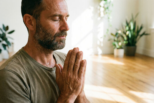 Authentic mindful practice: Sweaty middle-aged man with closed eyes and clasped hands meditating in sunny room. Men's health, inner peace and self-care routine