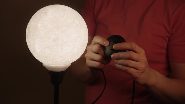 Unrecognizable person using dimmer switch to adjust light intensity of spherical lamp shaped like moon, changing ambiance of room from bright to dim, close-up, slow motion.