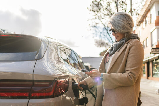 Woman charging electric car on urban street with smartphone
