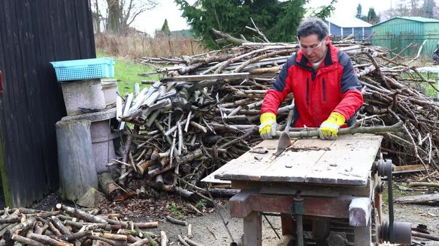active senior cutting branches on a circular saw as part of winter wood preparation, performing heavy manual work in a rural home setting with a pile of dry material 