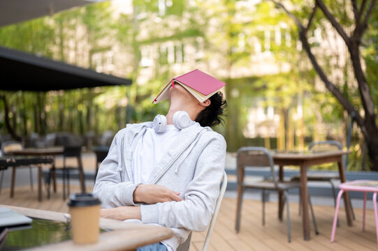 Young man putting a book covering face feeling tired or sleepy while sitting at table in garden cafe