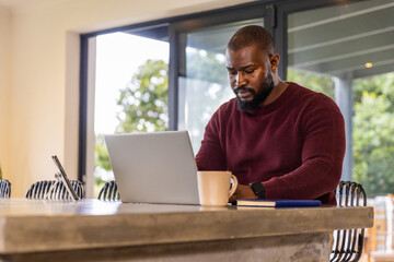 African American man sitting at counter in maroon sweater working on laptop with mug, copy space © wavebreak3