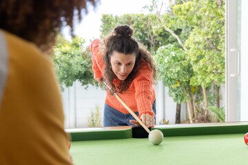 Naklejka premium Female friends playing on pool table at home with woman in orange sweater aiming wooden cue