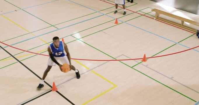 African american man in gym picking up ball starting orange cone dribble as teammate stepping up