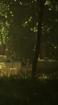 Tables and chairs are arranged in an outdoor space. Sunlight filters through the trees, creating a bright scene. Plants surround the area, adding green elements to the setting.